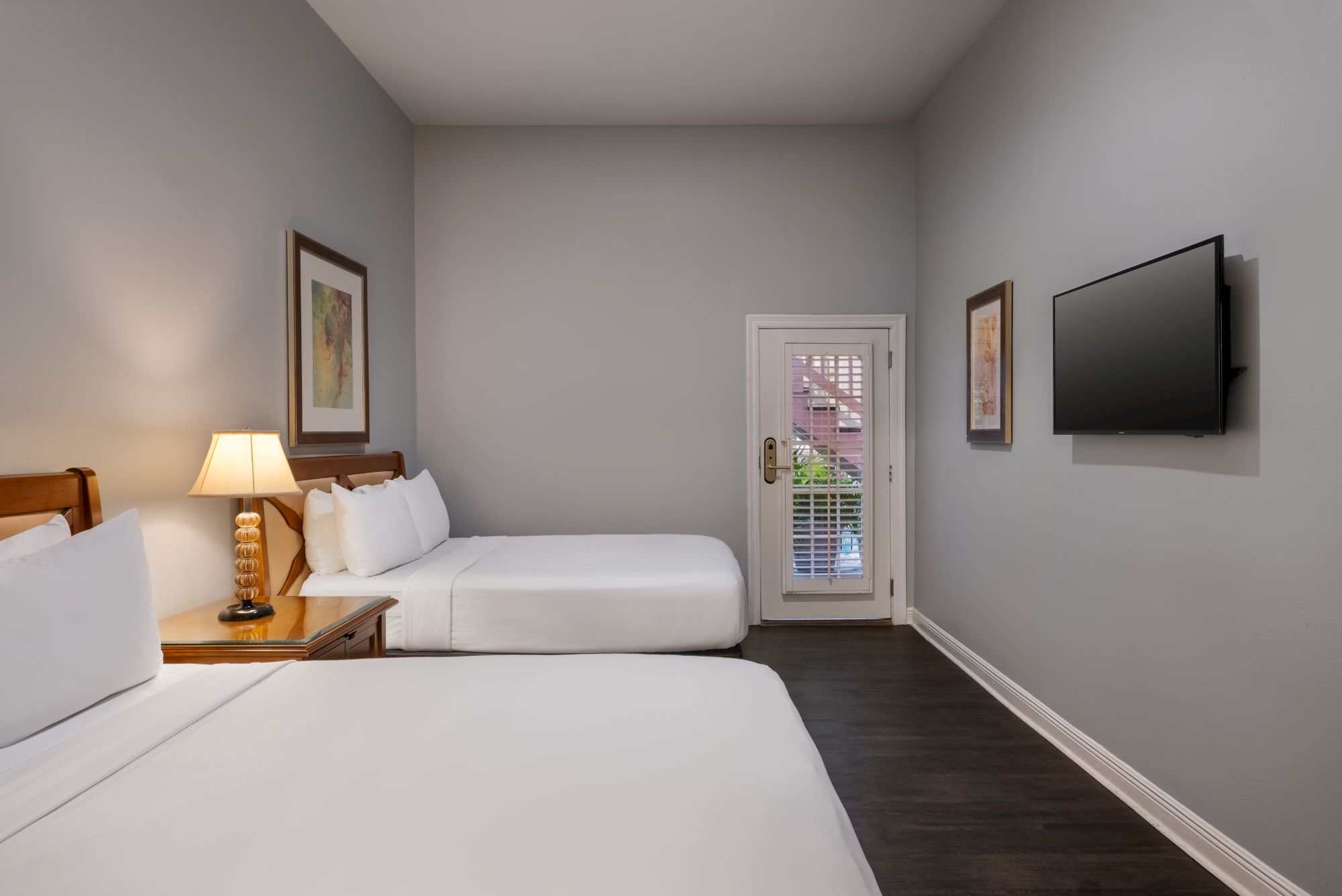 The image shows a hotel room with two beds, a TV, wall art, a lamp, and a door leading outside, with a minimalist decor style.