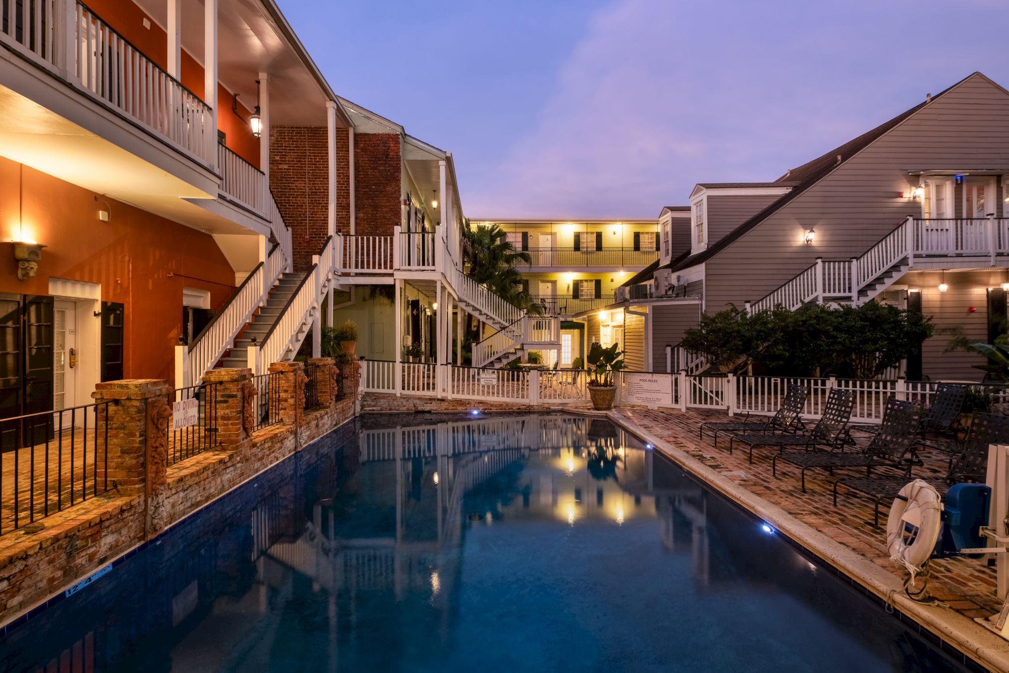 A serene outdoor pool surrounded by multi-story buildings with white railings, illuminated under the evening sky.