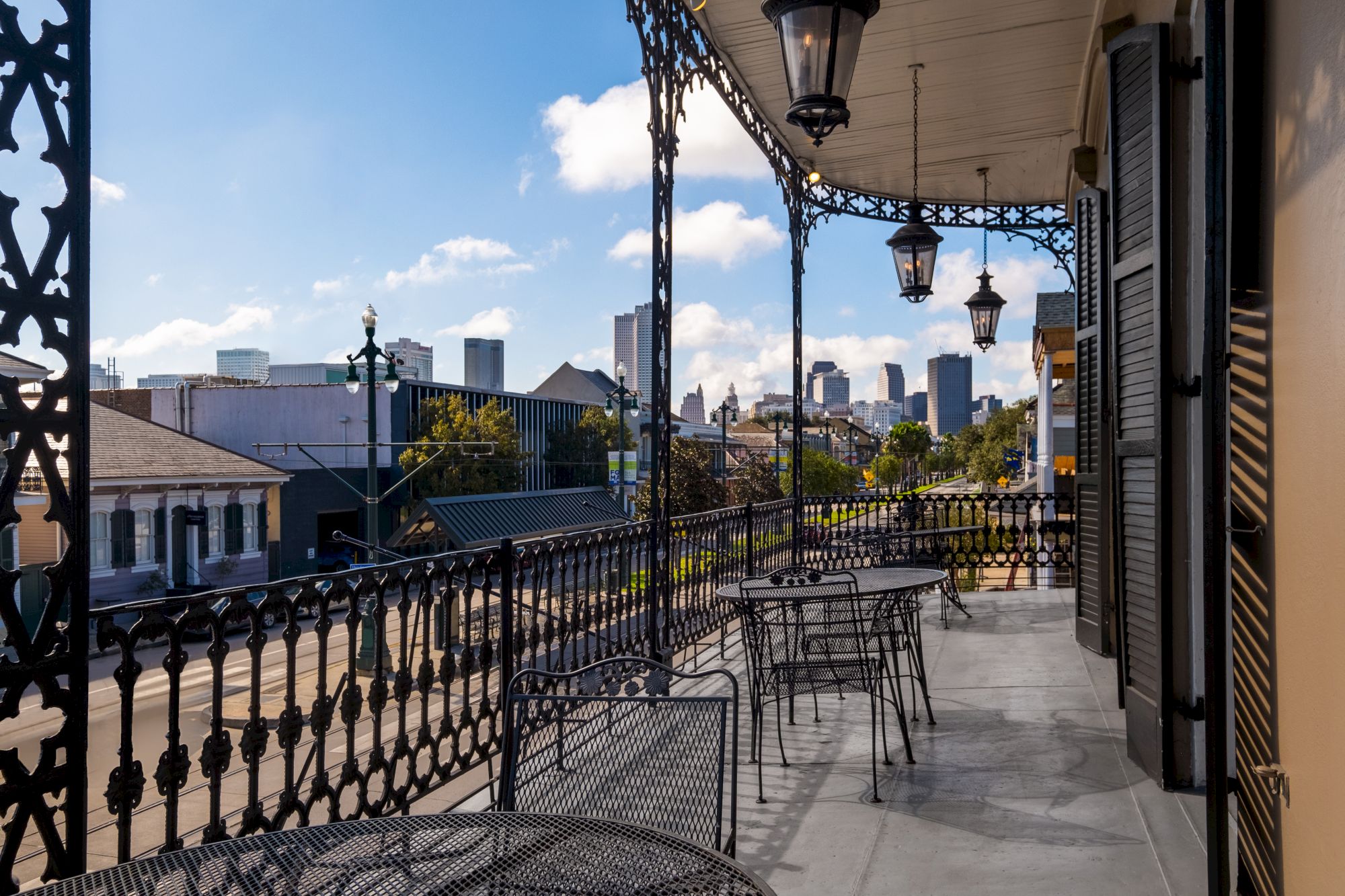 A balcony with ornate railings overlooks a street. Tables and chairs are set up, with a city skyline visible in the background.