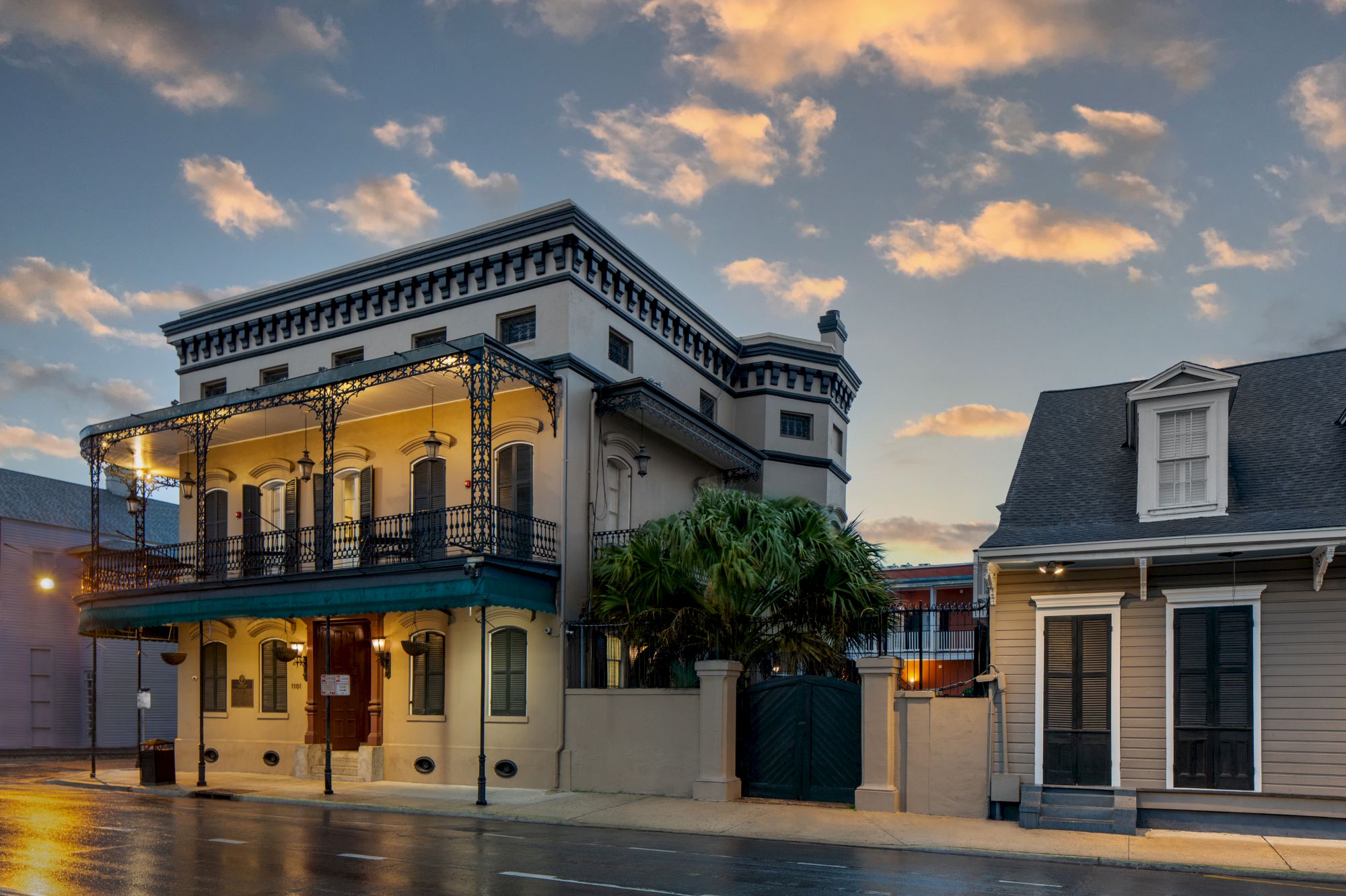 A historic, multi-story building with ornate ironwork balconies stands beside a smaller house, set against a sunset sky.