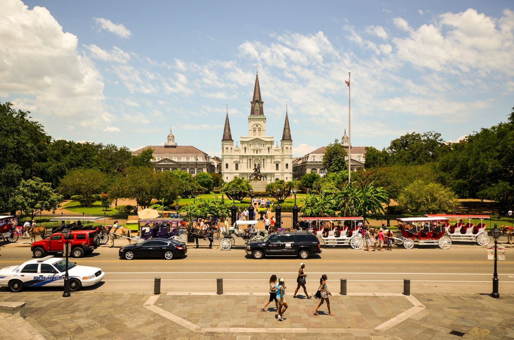 The image shows a bustling square with a historic building, lush gardens, and people walking and riding horse-drawn carriages.