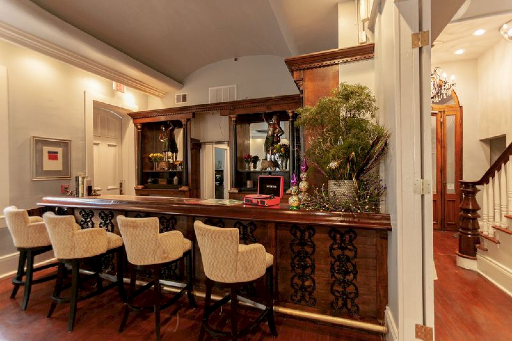 This image shows a cozy bar area with four upholstered chairs, ornate wooden counter, decorative plants, and a staircase in the background.