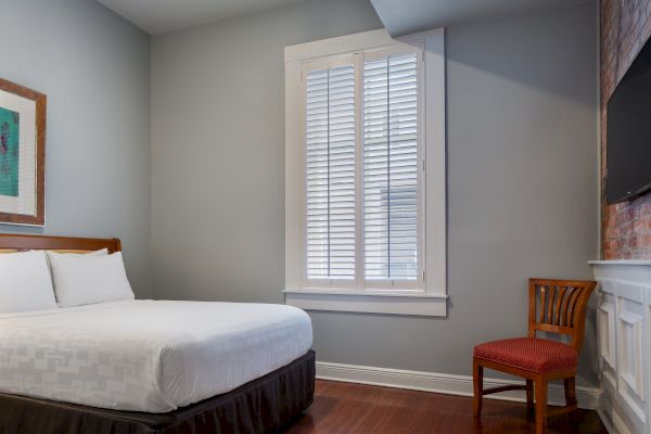 A simple room with a bed, chair, window, and wall art in neutral tones and a wooden floor.