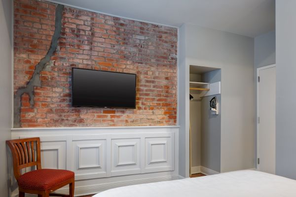 A room with a brick accent wall, mounted TV, wooden chair, and a partially visible bed, featuring a closet with hangers to the right.