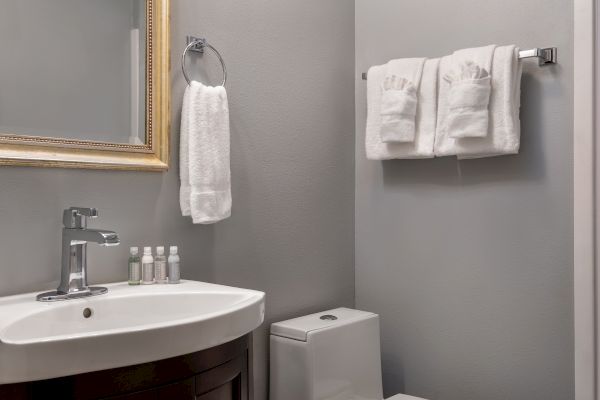 A modern bathroom with a sink, a mirror, toiletries, white towels on the rack, and a toilet against gray walls.