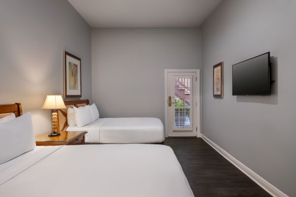 The image shows a hotel room with two beds, a nightstand with a lamp, wall art, a flat-screen TV, and a door leading outside.