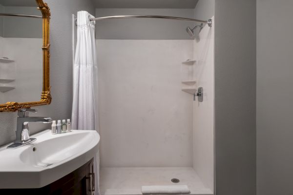 The image shows a bathroom with a shower, white curtain, sink, mirror, and toiletries on the counter.