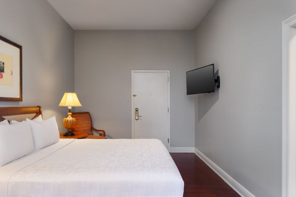 A neatly arranged hotel room with a bed, wall-mounted TV, door, lamp, and chair. Light gray walls and wood flooring complete the look.