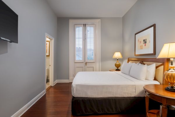 The image shows a cozy bedroom with a neatly made bed, lamps, framed artwork, wooden flooring, and a wall-mounted TV.