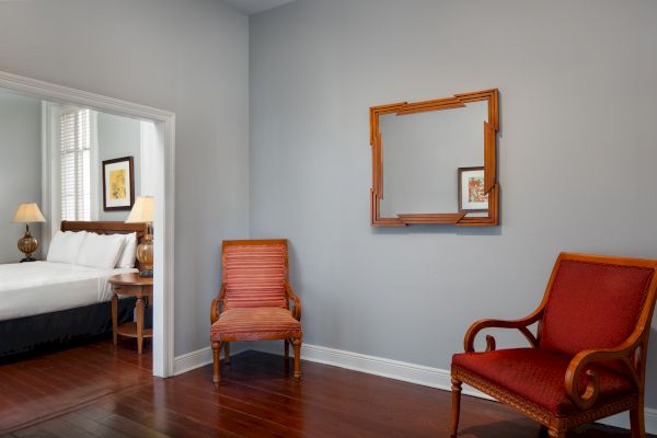 A room with two wooden chairs and a decorative frame on the wall, leading to a bedroom with a bed and nightstand visible.