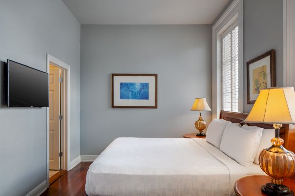 A cozy bedroom with a bed, two lamps, wall art, and a TV. Walls are light gray, enhancing the calm ambiance of the room.