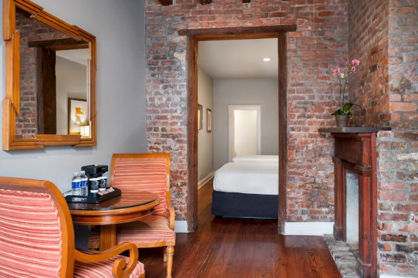 A cozy room with brick walls, a small table with chairs, a mirror, and a view into a bedroom with a bed and fireplace.