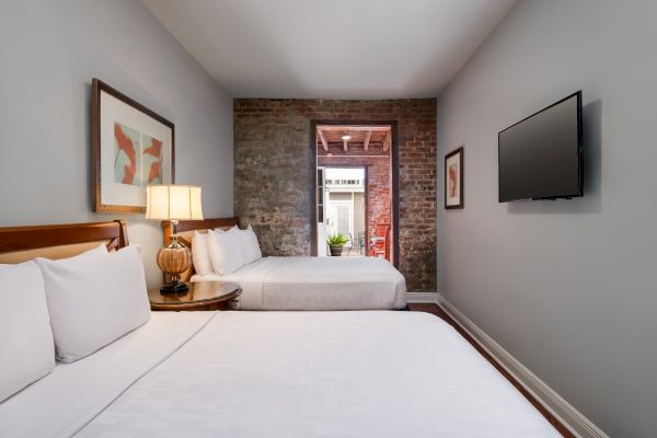 A room with two beds, framed art, a lamp, a TV, and an exposed brick wall, leading to a patio with plants visible through a doorway.