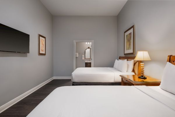 The image shows a hotel room with two beds, a flat-screen TV, wall art, and a lamp on a wooden nightstand next to one bed.