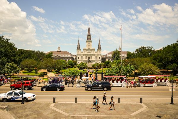 The image shows a crowded street with carriages in front of a historic building and park on a sunny day.