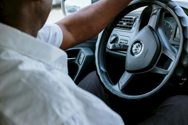 A person is sitting in a car, holding the steering wheel with one hand while looking out the window. The car interior is partially visible.