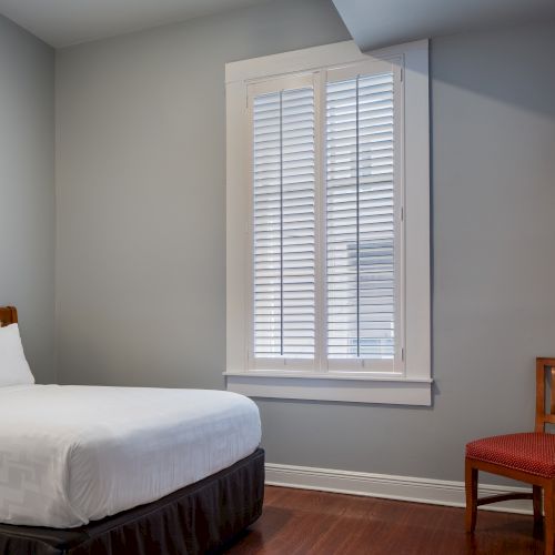 The image shows a neatly arranged bedroom with a bed, a wooden chair, a window with blinds, and a wall-mounted TV.