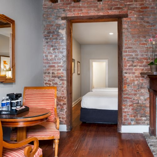 A cozy room with brick walls, a fireplace, a round table with two chairs, a mirror, and a view into a bedroom with a made bed.