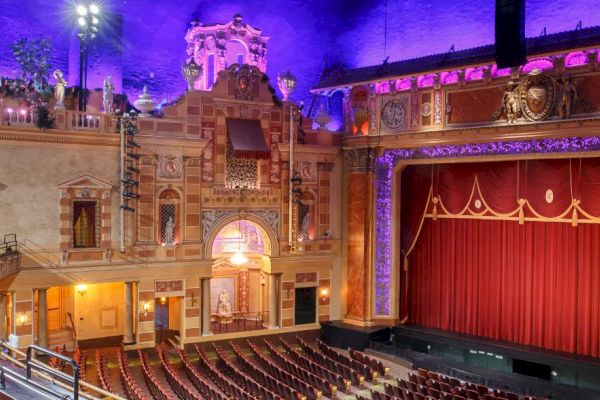 An ornate theater interior with balconies, red seating, and a grand stage with a red curtain, illuminated by colorful lights.