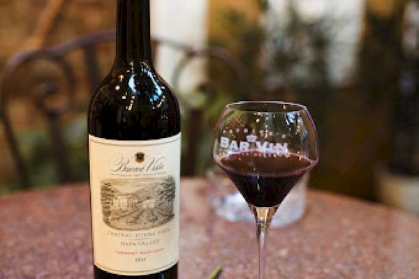 A wine bottle, filled glass, red rose, and a blue napkin on a table in a cozy setting.