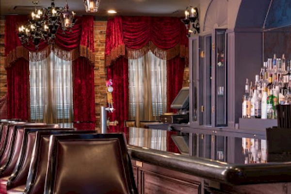 The image shows an elegant bar with leather stools, hanging lights, red curtains, and a well-stocked drink shelf, offering a cozy atmosphere.