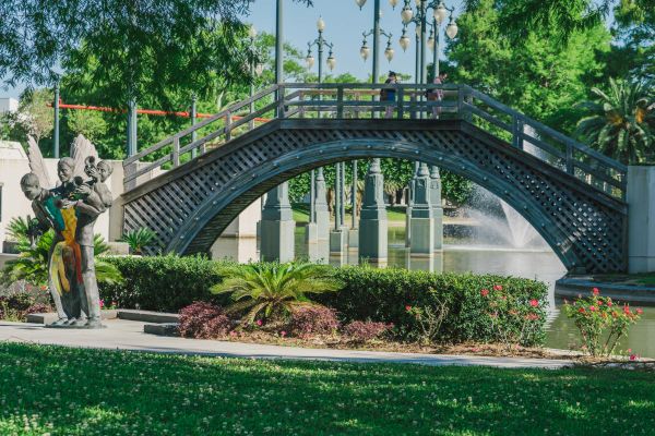 A scenic park features a wooden bridge over a pond, a fountain, green plants, and a modern sculpture amidst lush greenery and trees.