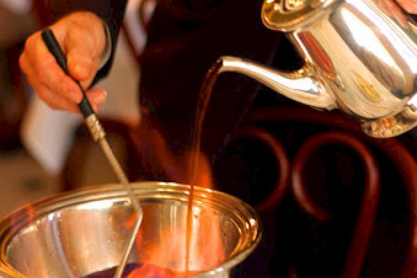 A person is preparing a flaming beverage by pouring liquid from a silver teapot into a bowl with fire at a restaurant table.