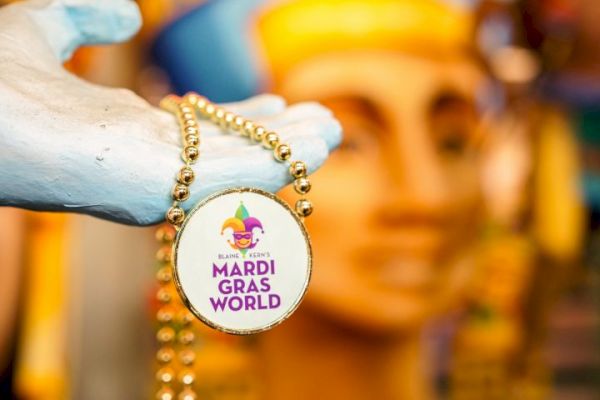 A hand holds a beaded necklace with a "Mardi Gras World" medallion, featuring a jester hat logo, against a colorful background.