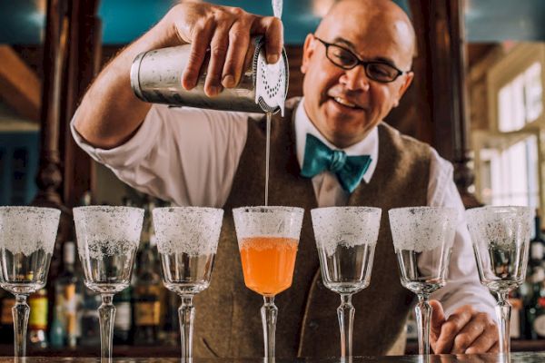 A bartender pours an orange cocktail into one of six rimmed glasses while wearing a vest and bow tie.