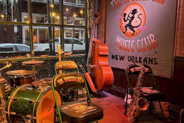 A jazz club stage with drums, a double bass, and a saxophone; banner reads "The Spotted Cat Music Club, New Orleans."