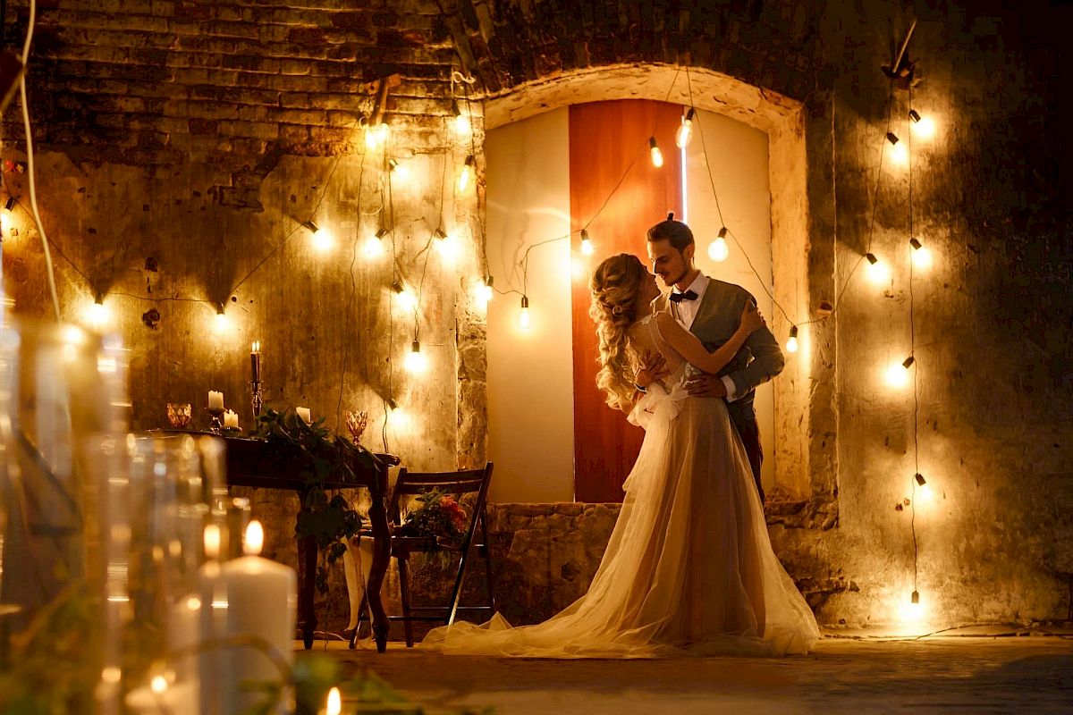 A couple is dancing in a dimly lit, rustic setting decorated with string lights and candles.